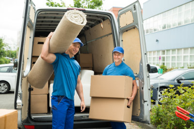 Two Male Workers Carrying Carpet And Cardboard Boxes In Front Of Van